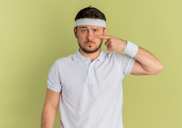 Young fitness man in white shirt with headband pointing with finger to his eye standing over olive wall