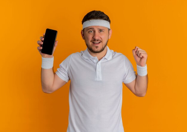 オレンジ色の壁の上に立って幸せで前向きなスマートフォンを握りこぶしを示すヘッドバンドと白いシャツの若いフィットネス男