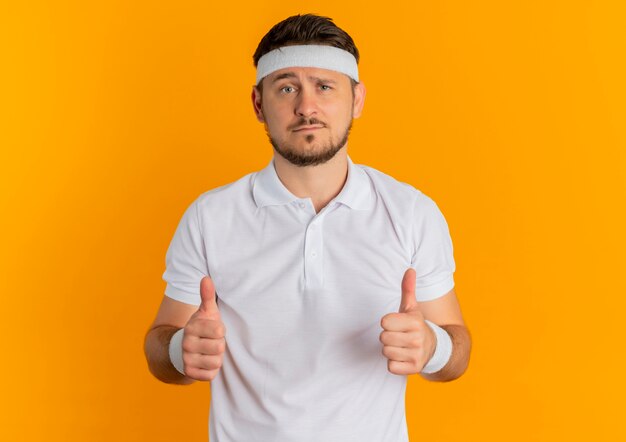 Young fitness man in white shirt with headband showing thumbs up looking to the front standing over orange wall