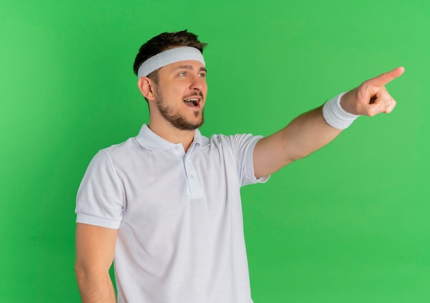 Giovane uomo di forma fisica in camicia bianca con fascia sorridendo allegramente puntando il dito a qualcosa in piedi sopra la parete verde