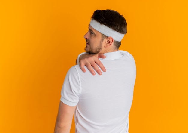 Young fitness man in white shirt with headband standing with his back touching his shoulder having pain over orange wall