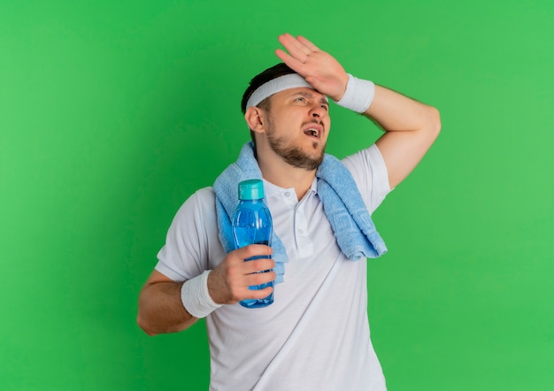 Young fitness man in white shirt with headband and towel around his neck holding bottle of water tired and exhausted after workout standing over green background