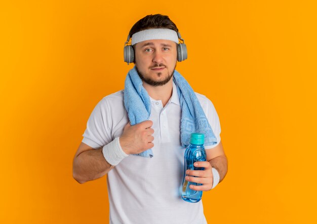 Молодой фитнес-мужчина в белой рубашке с повязкой на голову и полотенцем на шее держит бутылку воды, глядя в камеру с уверенным выражением лица, стоя на оранжевом фоне
