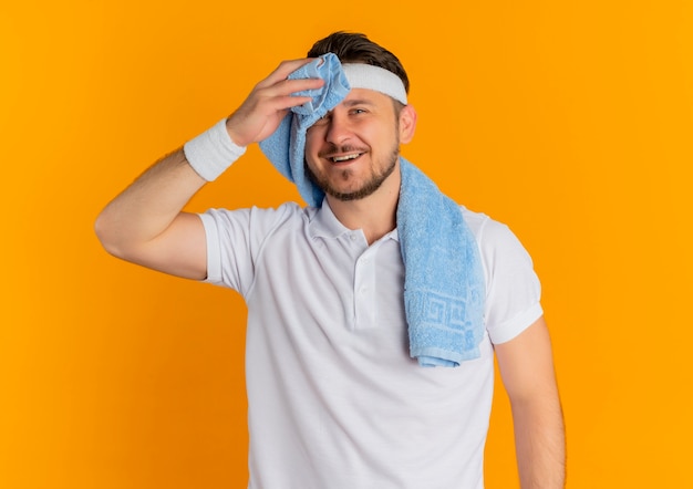 Young fitness man in white shirt with headband and towel around neck holding looking at camera tired and smiling standing over orange background