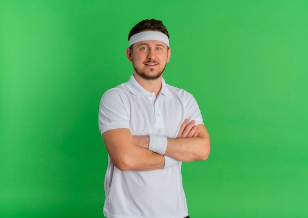 Young fitness man in white shirt with headband with crossed hands on chest looking to the front with confident expression standing over green wall