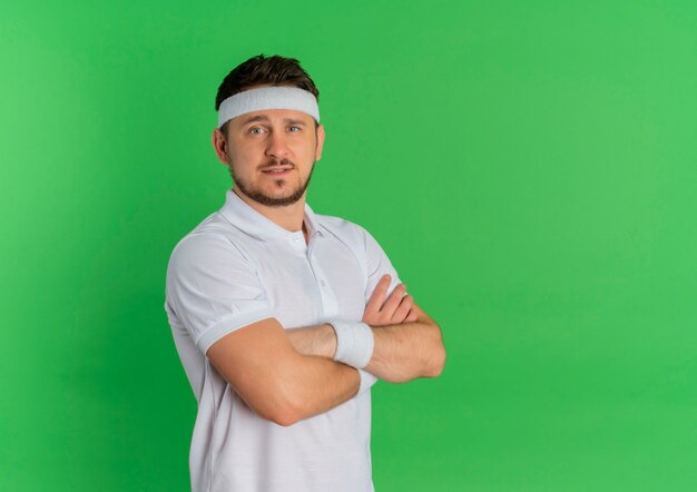 Young fitness man in white shirt with headband with crossed hands on chest looking to the front with confident expression standing over green wall