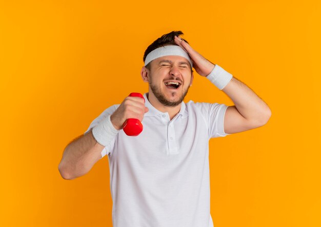 Young fitness man in white shirt with headband working out with dumbbell looking confused, forgot standing over orange wall