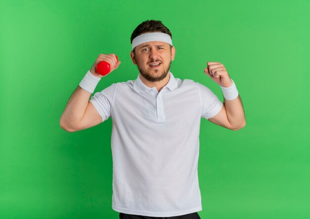 Young fitness man in white shirt with headband working out with dumbbell strained and confident standing over green wall