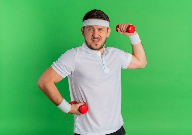 Молодой фитнес-мужчина в белой рубашке с повязкой на голове тренируется с напряженными и уверенными гантелями, стоя у зеленой стены