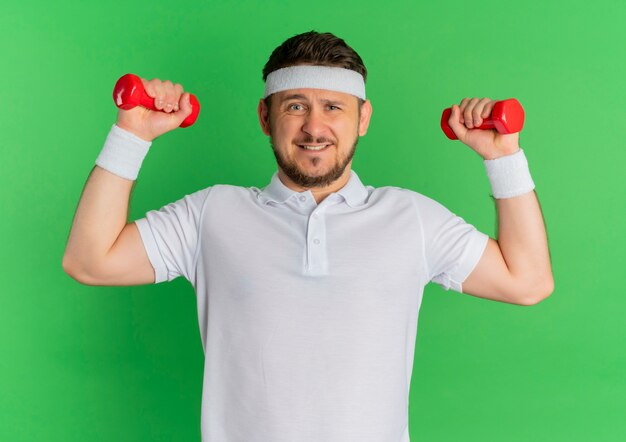 Young fitness man in white shirt with headband working out with dumbbells strained and confident standing over green wall