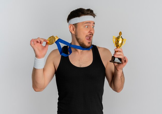 Young fitness man with headband and gold medal around his neck holding trophy looking amazed and surprised standing over white background