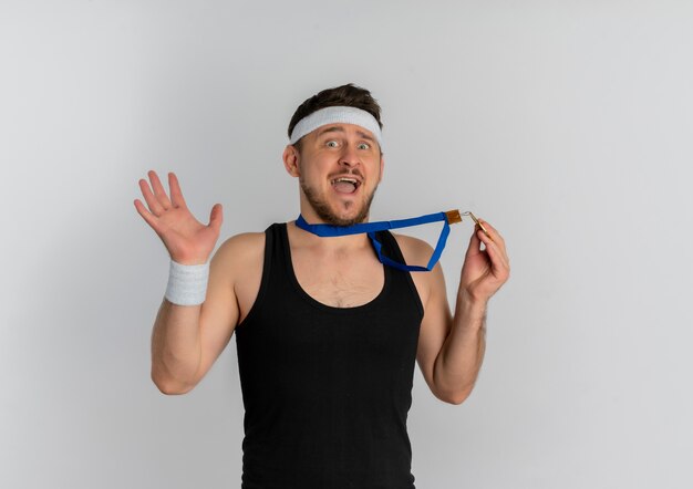 Young fitness man with headband and gold medal around his neck looking at camera surprised and amazed standing over white background