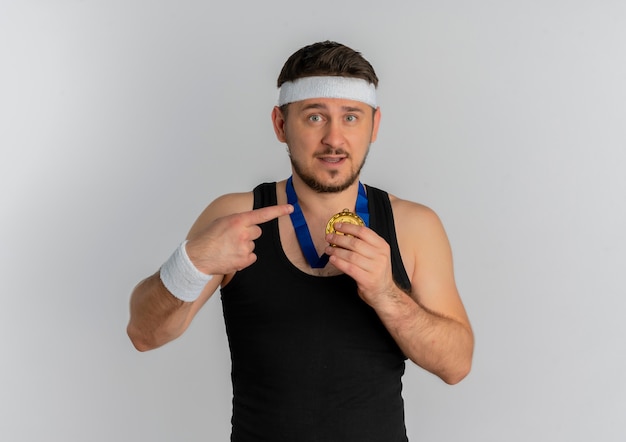 Молодой фитнес-мужчина с повязкой на голову и золотой медалью на шее, указывая пальцем на нее, выглядит уверенно, стоя на белом фоне