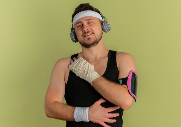 Young fitness man with headband and headphones looking at camera training with smartphone armband standing over olive background