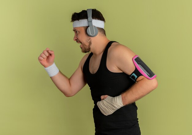 Молодой фитнес-мужчина с повязкой на голову и наушниками тренируется с повязкой для смартфона, стоящей на оливковом фоне