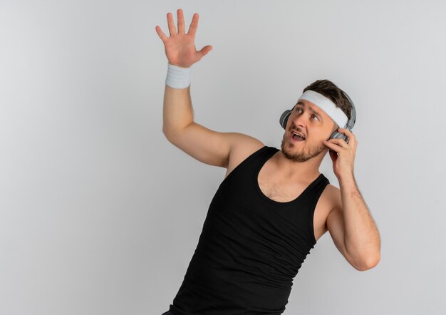 Young fitness man with headband and headpones looking aside emotional and excited standing over white background