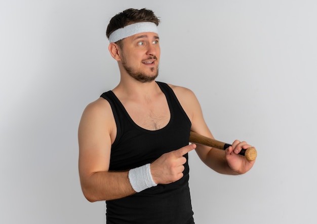 Young fitness man with headband holding baseball bat looking aside confused standing over white background