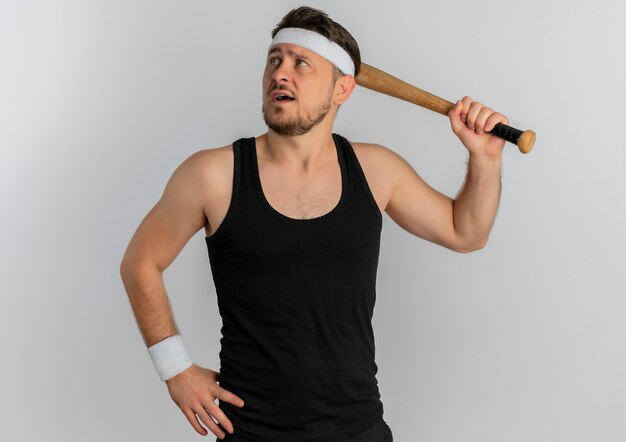 Young fitness man with headband holding baseball bat looking aside with confident expression standing over white background