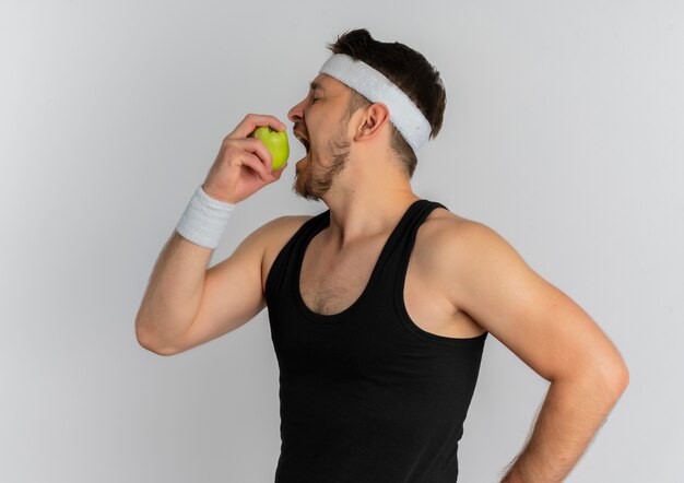 Young fitness man with headband holding green apple biting it standing over white background