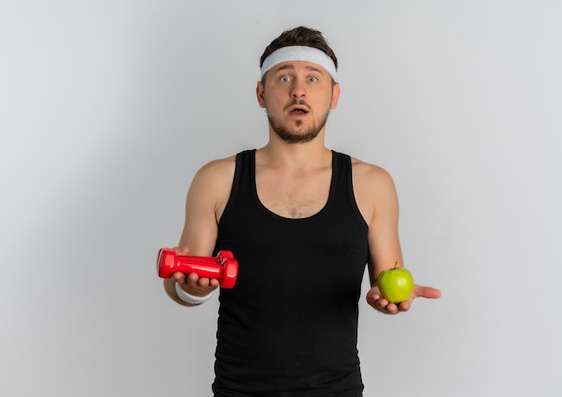 Молодой фитнес-мужчина с повязкой на голову, держащий зеленое яблоко и гантель, глядя в камеру, смущается, стоя на белом фоне