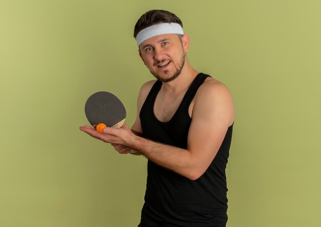 Молодой фитнес-мужчина с повязкой на голову, держащей ракетку и мячи для настольного тенниса, собирается играть, улыбаясь, стоя на оливковом фоне