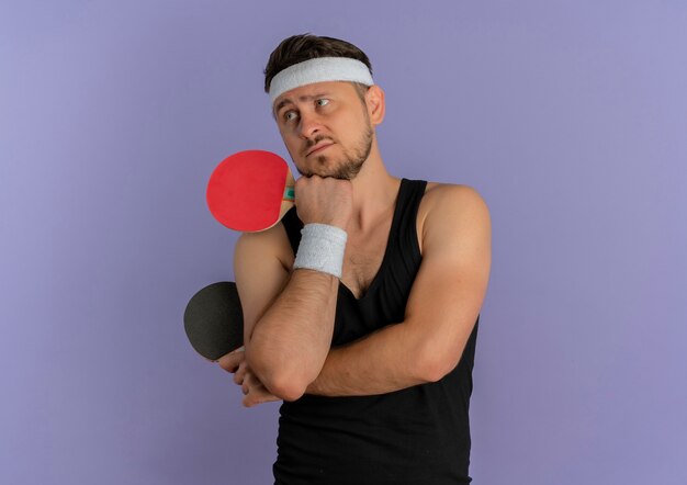 Young fitness man with headband holding two rackets for table tennis looking aside with pensive expression standing over purple wall