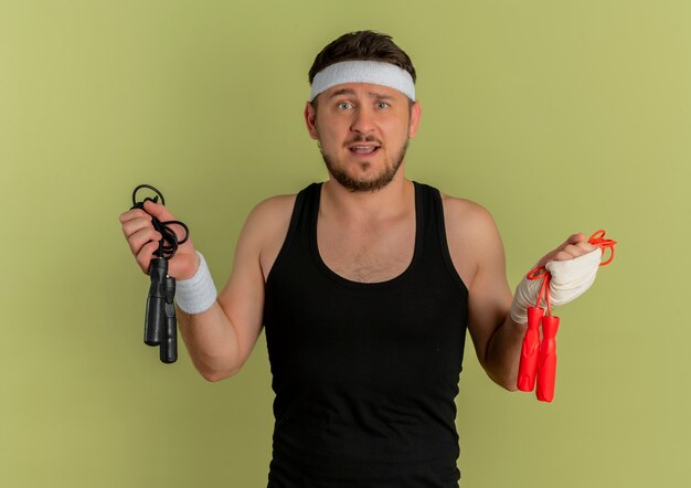 young fitness man with headband holding two skipping ropes looking confused and uncertain having doubts standing over olive background