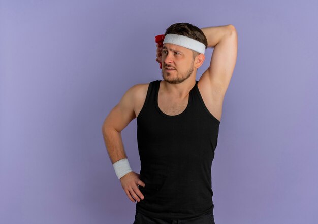 Young fitness man with headband looking aside doing exercises with dumbbell standing over purple wall