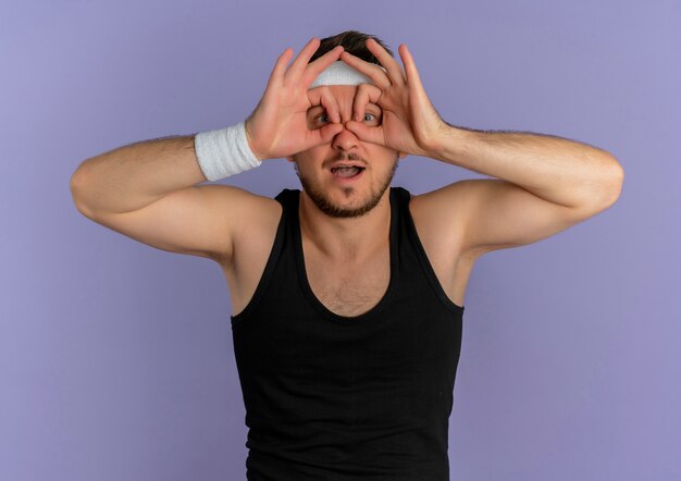 Young fitness man with headband making binocular gesture with fingers looking to the front through fingers standing over purple wall