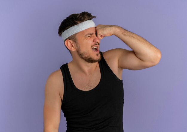Young fitness man with headband punching himself looking confused standing over purple wall