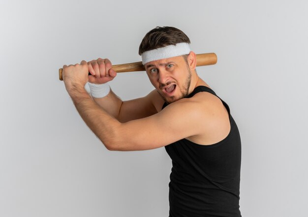 Young fitness man with headband swinging a baseball bat emotional and excited standing over white background