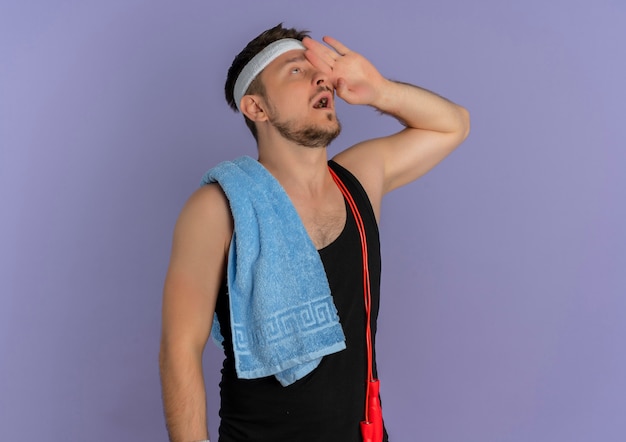 Young fitness man with headband and towel on shoulder looking up with hand over head standing over purple wall