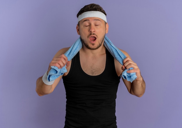 Free photo young fitness man with headband and towel on shoulder with closed eyea and wide open mouth standing over purple wall