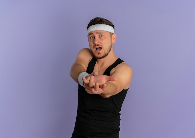 Молодой фитнес-мужчина с повязкой на голову, тренирующийся, протягивая руки, выглядел удивленным, стоя у фиолетовой стены