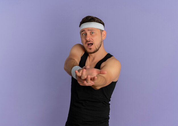 Young fitness man with headband working out stretching arms out looking surprised standing over purple wall