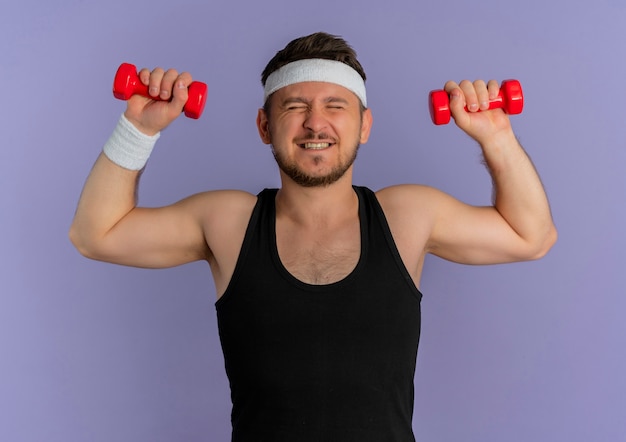 Giovane uomo fitness con archetto lavorando con manubri guardando fiducioso e teso in piedi su sfondo viola