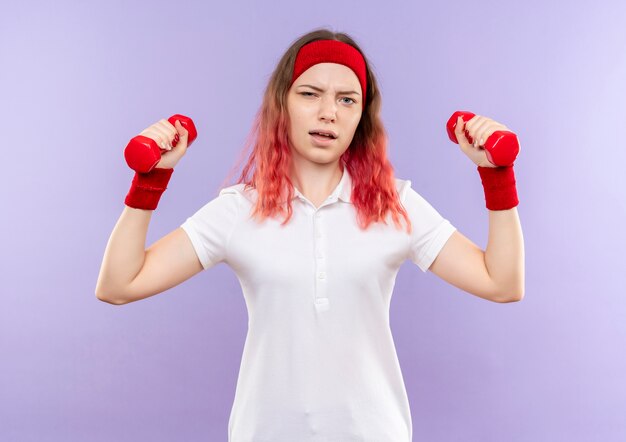 Young fitness woman doing exercises using dumbbells with serious face standing over purple wall