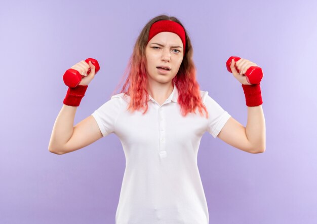 Молодая фитнес-женщина делает упражнения с гантелями с серьезным лицом, стоящим над фиолетовой стеной