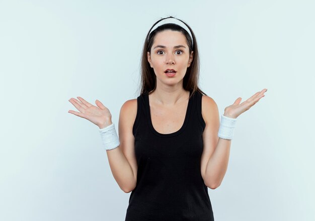 young fitness woman in headband  confused and uncertain spreading arms to the side standing over white wall
