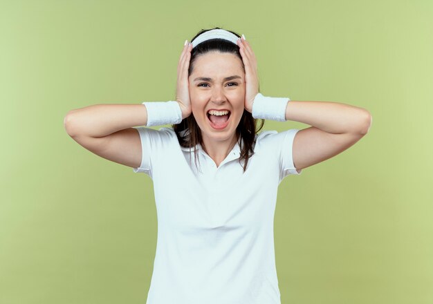 Young fitness woman in headband crazy mad shouting closing her ears with hands standing over light background