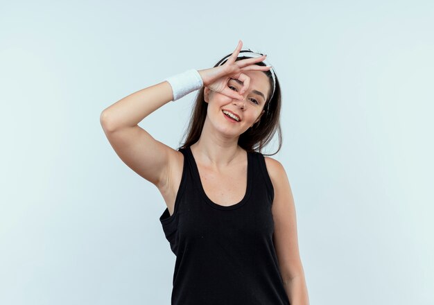 Young fitness woman in headband doing ok sign with fingers looking at camera through this sign smiling standing over white background