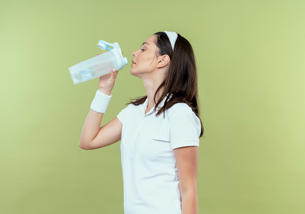 Free photo young fitness woman in headband drinking water after workout standing over light background