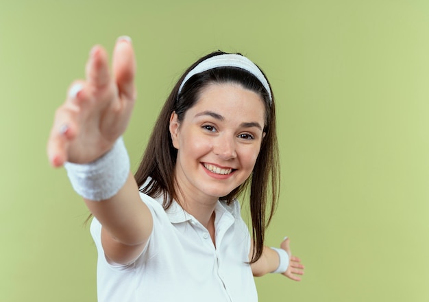 молодая фитнес-женщина в повязке на голову счастлива и позитивна, делая приветственный жест руками, стоящими над светлой стеной