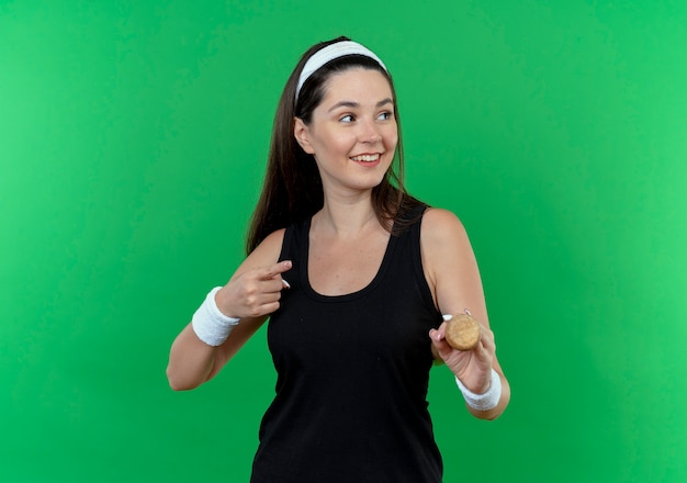 Молодая фитнес-женщина в повязке на голову, держащая бейсбольную биту, глядя в сторону, улыбаясь со счастливым лицом, указывая пальцем в сторону, стоя на зеленом фоне