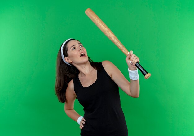 Young fitness woman in headband holding baseball bat looking at it confused standing over green background