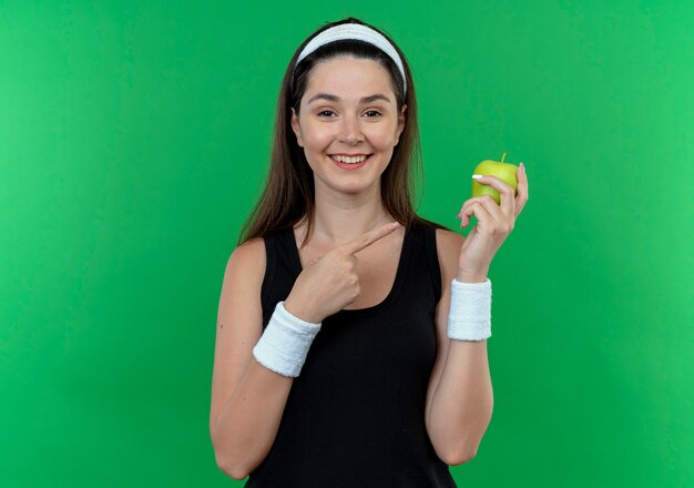 녹색 배경 위에 유쾌하게 서 웃고 그것에 손가락으로 가리키는 녹색 사과를 들고 머리띠에 젊은 피트 니스 여자
