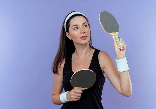 青い壁の上に立っている自信を持って表情を脇に見てテニステーブルのラケットを保持しているヘッドバンドの若いフィットネス女性