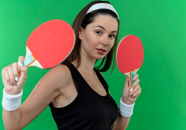 Young fitness woman in headband holding rackets for tennis table looking at camera with confident expression standing over green background