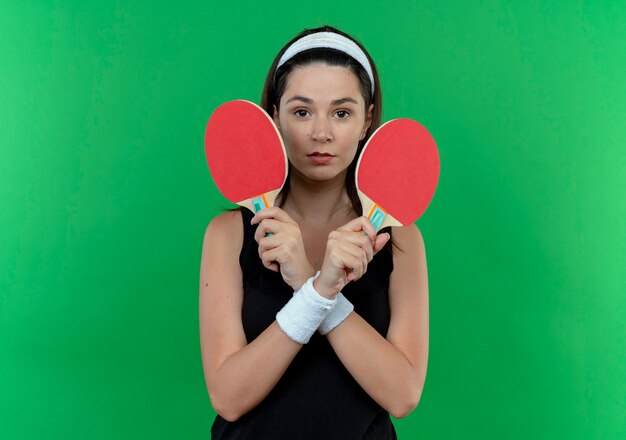 Молодая фитнес-женщина в головной повязке, держащая ракетки для теннисного стола, смотрит в камеру с серьезным лицом, скрещивающим руки, стоя на зеленом фоне