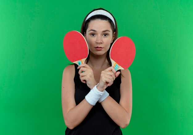 Young fitness woman in headband holding rackets for tennis table looking at camera with serious face crossing hands standing over green background